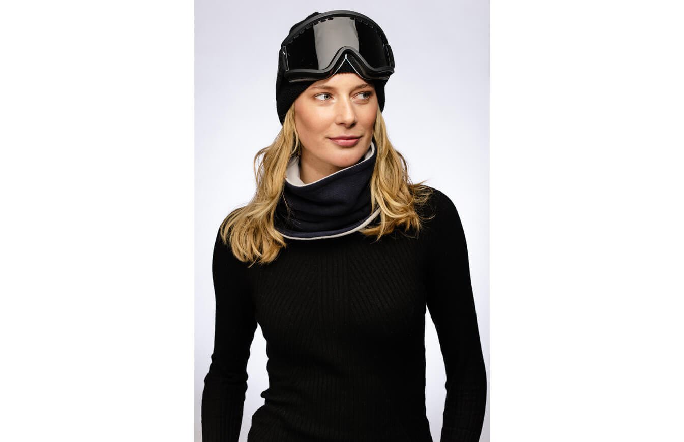 Frau mit schwarzem Pullover, Mütze und Skibrille trägt einen marineblauen Reversible Neckwarmer.