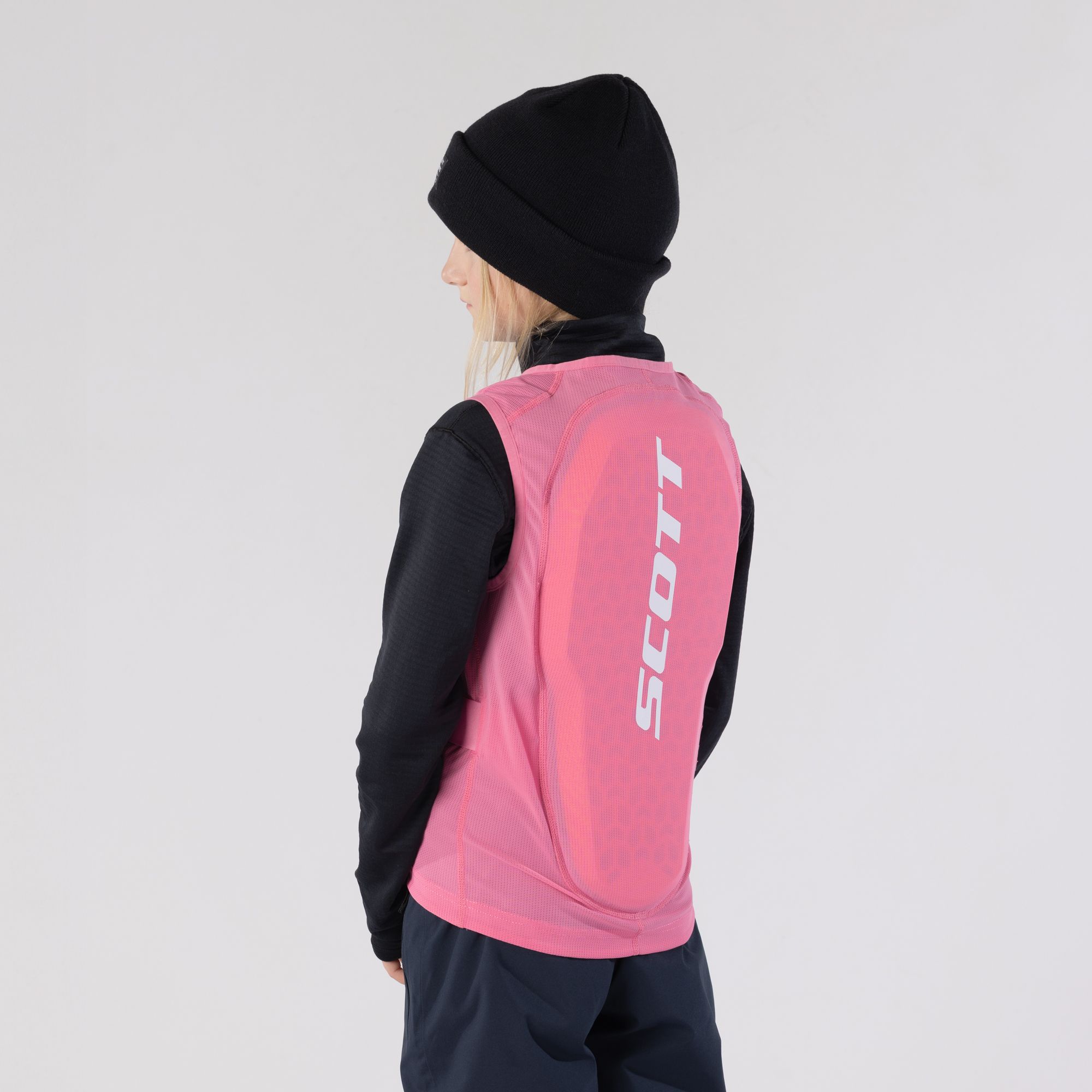 Ein junges Mädchen trägt eine schwarze Mütze, ein schwarzes Oberteil und eine pinkfarbene Weste mit dem Schriftzug "SCOTT" auf dem Rücken.