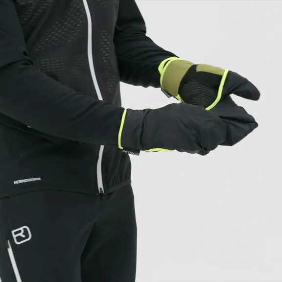 Una persona indossa i guanti Ortovox Fleece Grid Cover nei colori nero e verde. Il cappuccio di copertura è tirato sulle dita.