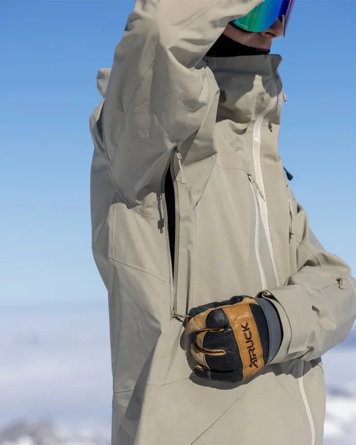 Una persona con occhiali e guanti da sci, che indossa una giacca beige con cerniera, davanti a un cielo blu e bianco sfocato.