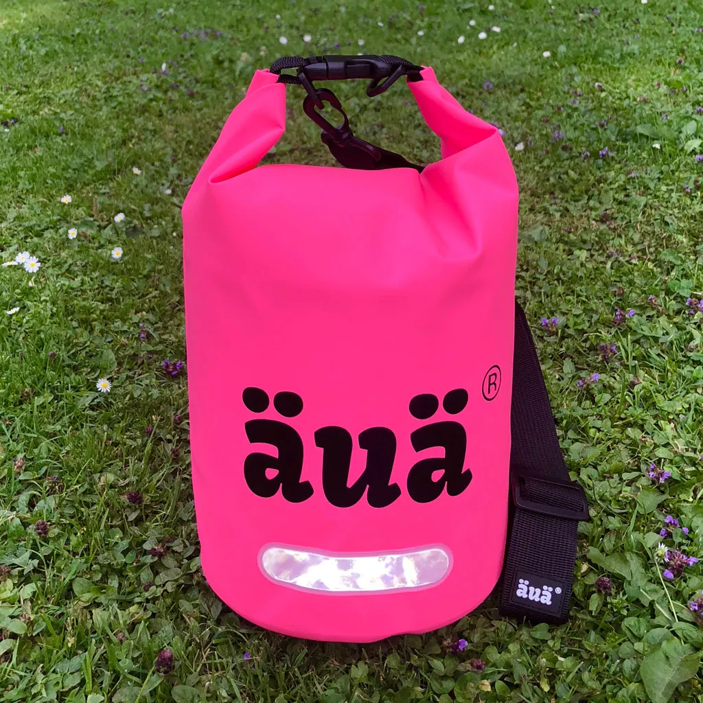 Pinker Dry Bag 5L auf grünem Rasen. Schwarze Tragegurte und reflektierendes Detail. "äua"-Logo in Schwarz.