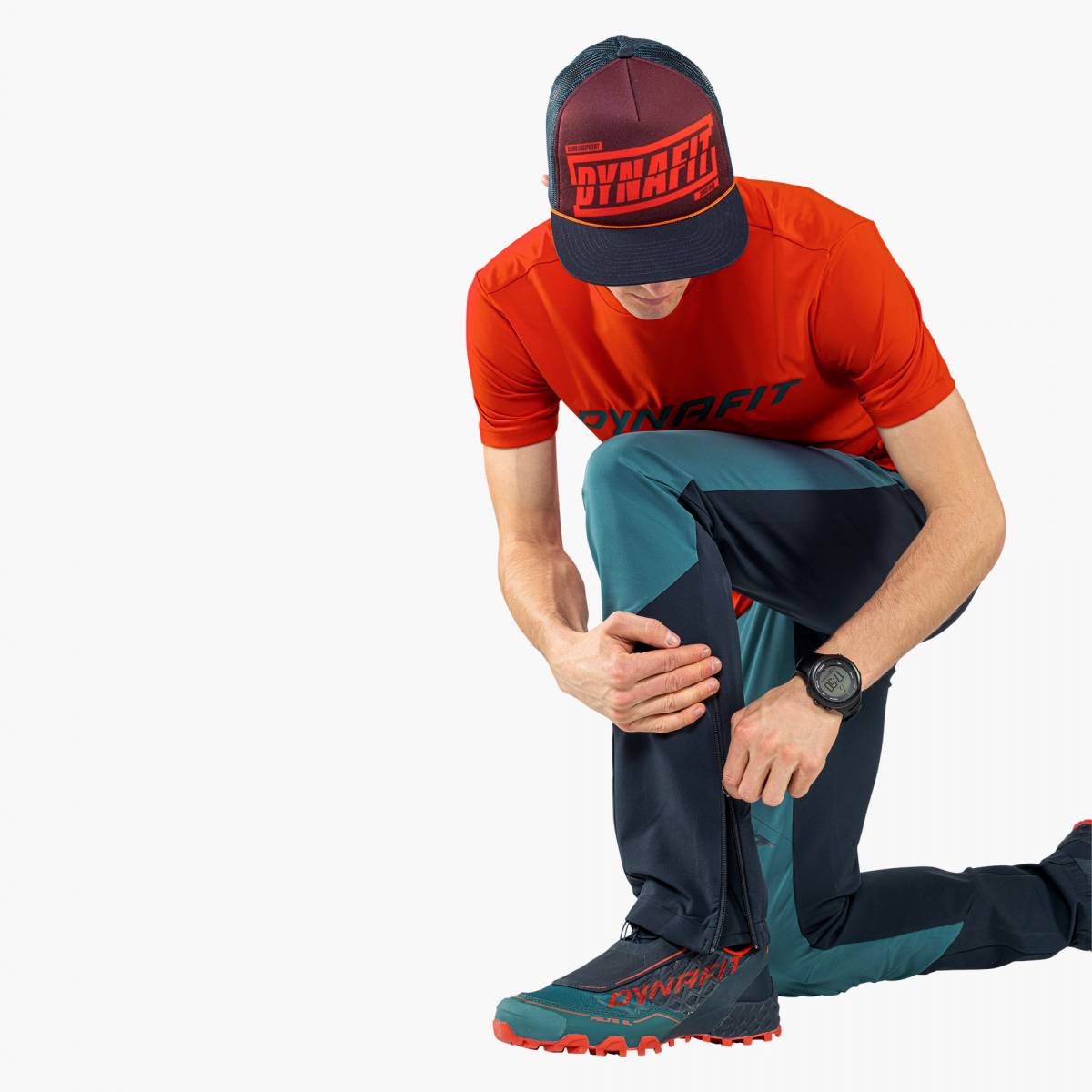 Man kneeling with Dynafit Graphic Trucker Cap and sportswear. View of the zipper of the pants.