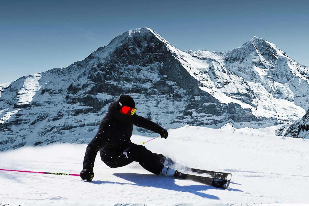 Skifahrer in schwarzer Kleidung, rote Brille, vor schneebedecktem Berg, blauer Himmel.