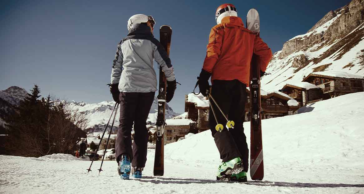 Zwei Skifahrer, mit Ski und Stöcken, von hinten in Winterkleidung, auf dem Schnee, im Hintergrund Berge und Häuser.