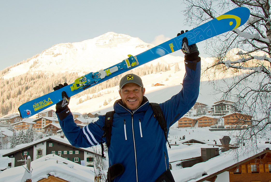 Ein Mann hält einen blauen Nebula Ski mit gelber Bindung über dem Kopf, im Hintergrund verschneite Berge und Häuser.