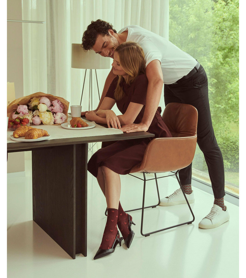 Paar am Tisch; Frau mit Velour Beat Socken Lipstick und Samtherzen, Mann umarmt sie.