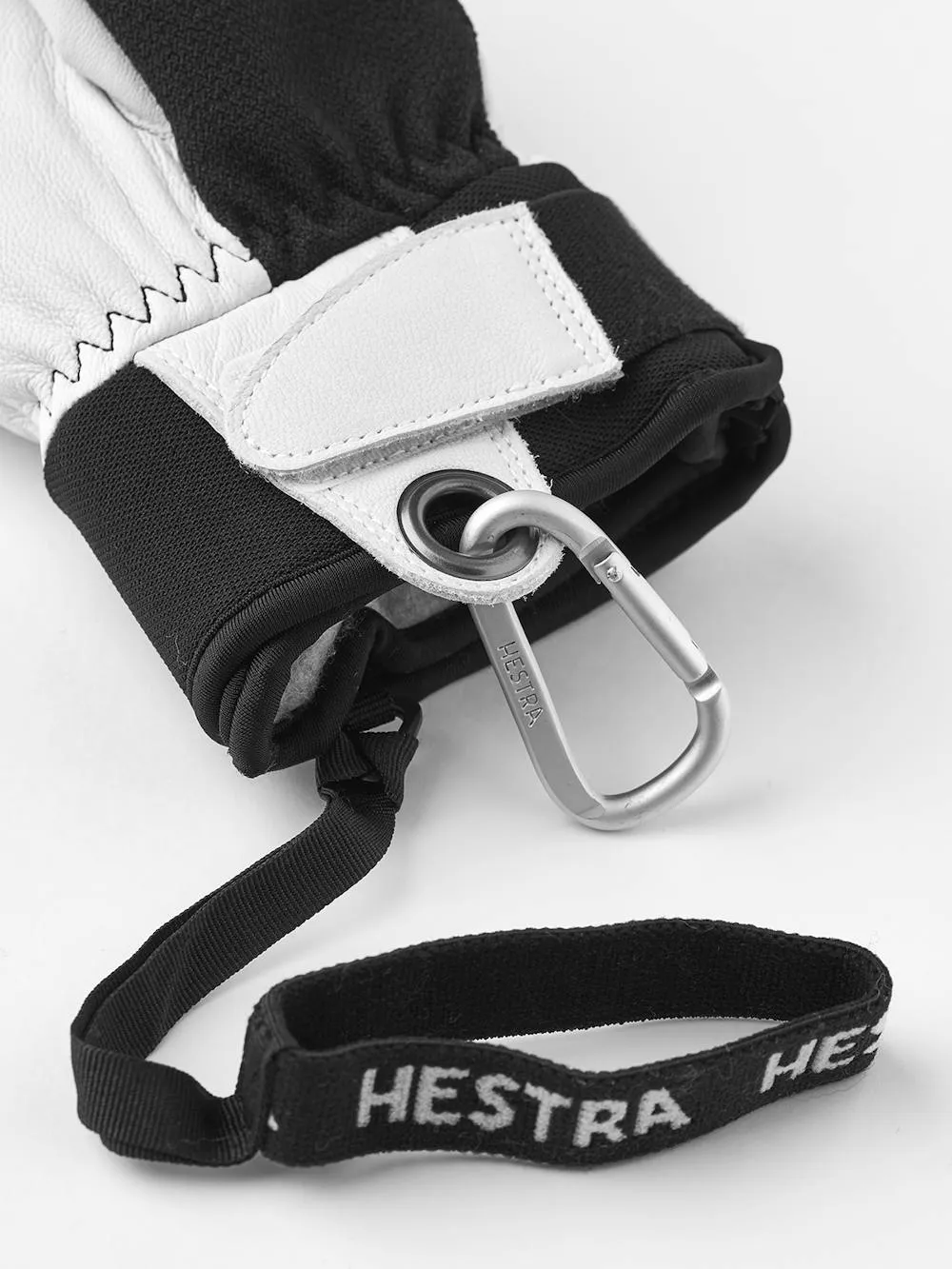 Primo piano di un guanto in bianco e nero con un moschettone e un nastro con la scritta "Hestra".