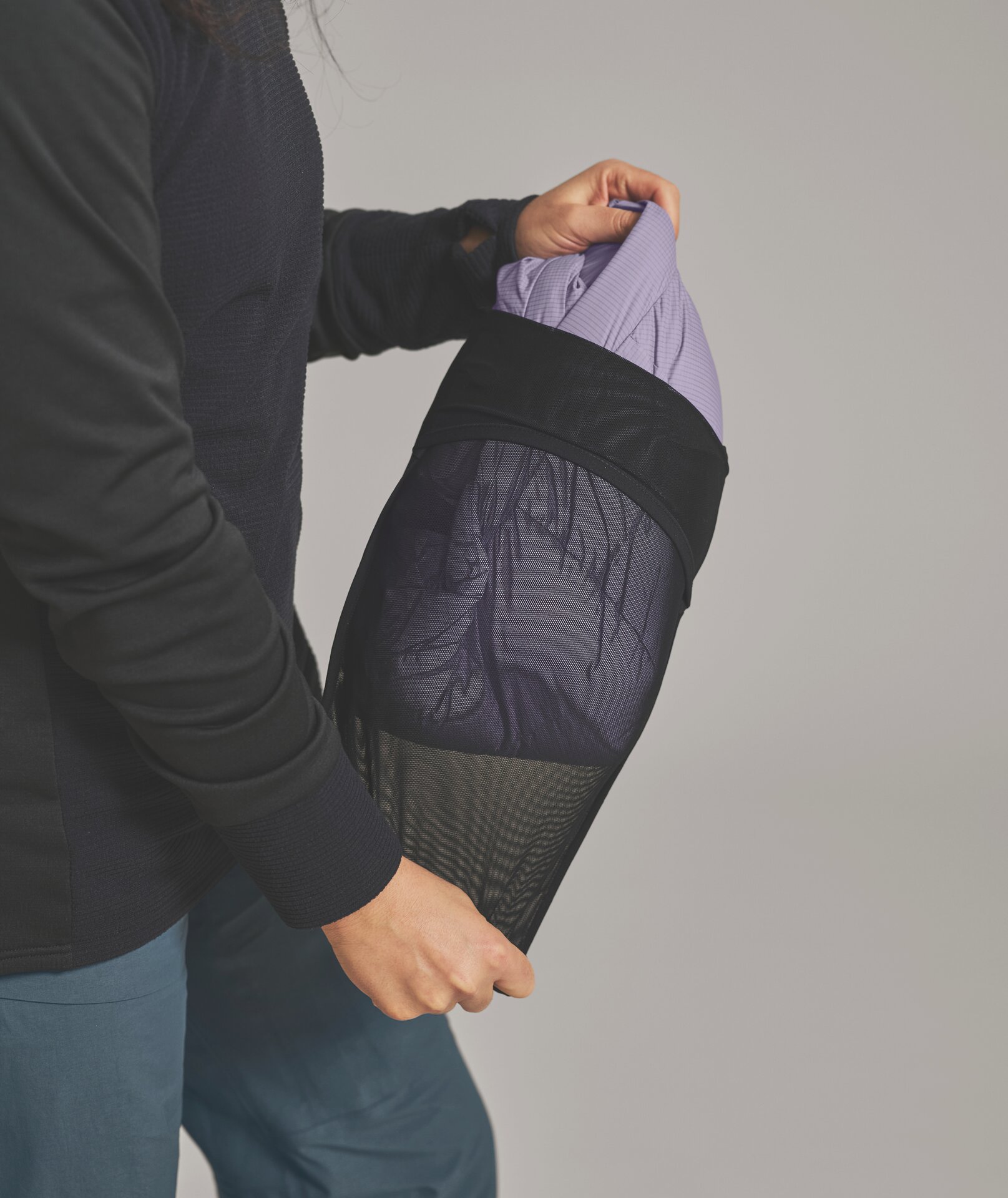Person packt lila Kleidung in einen schwarzen Netzbeutel. Die Person trägt einen schwarzen Pullover und grüne Hose.