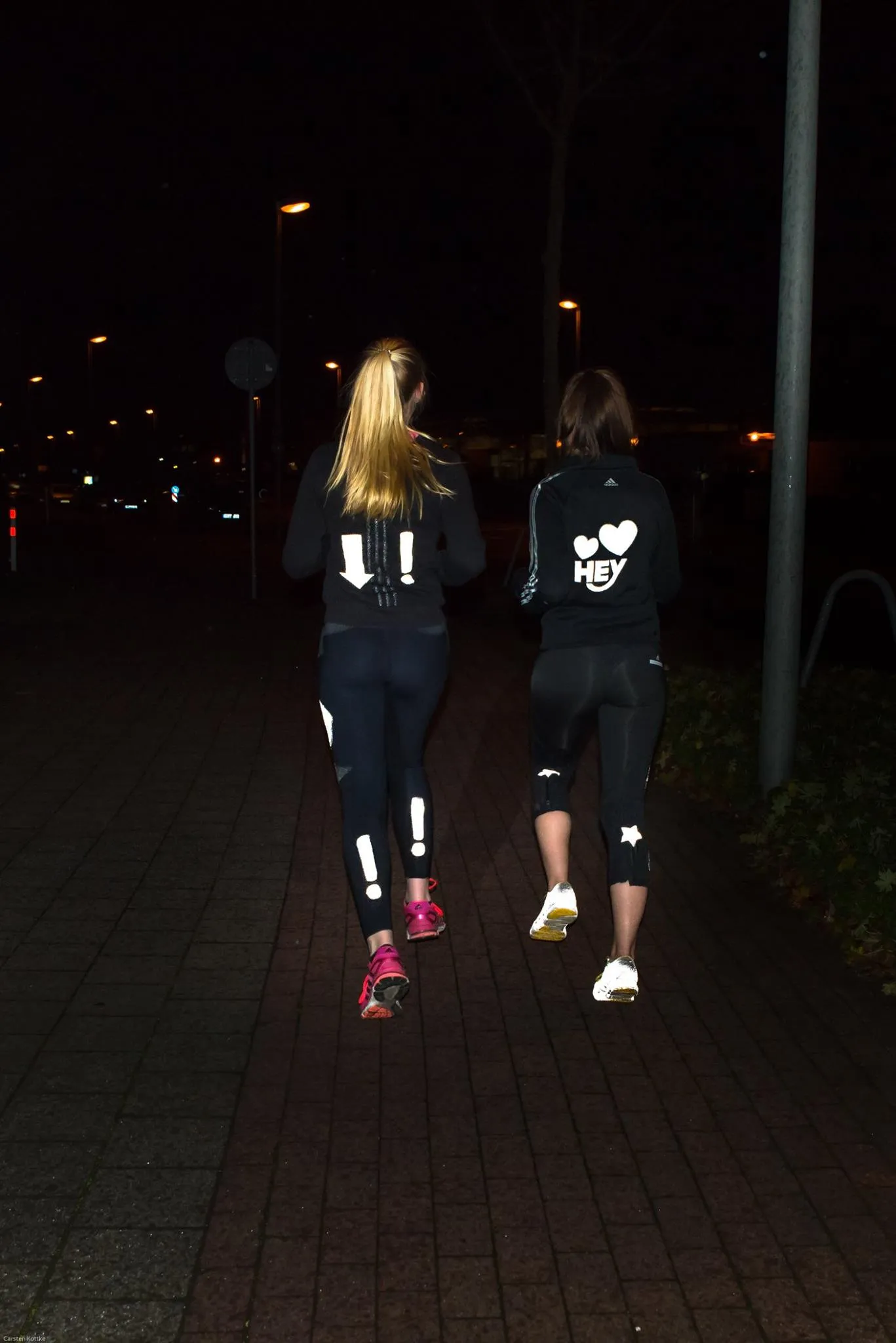 Zwei Frauen joggen nachts auf einem Pflasterweg, tragen reflektierende Sportkleidung. Straßenlaternen beleuchten die Dunkelheit.
