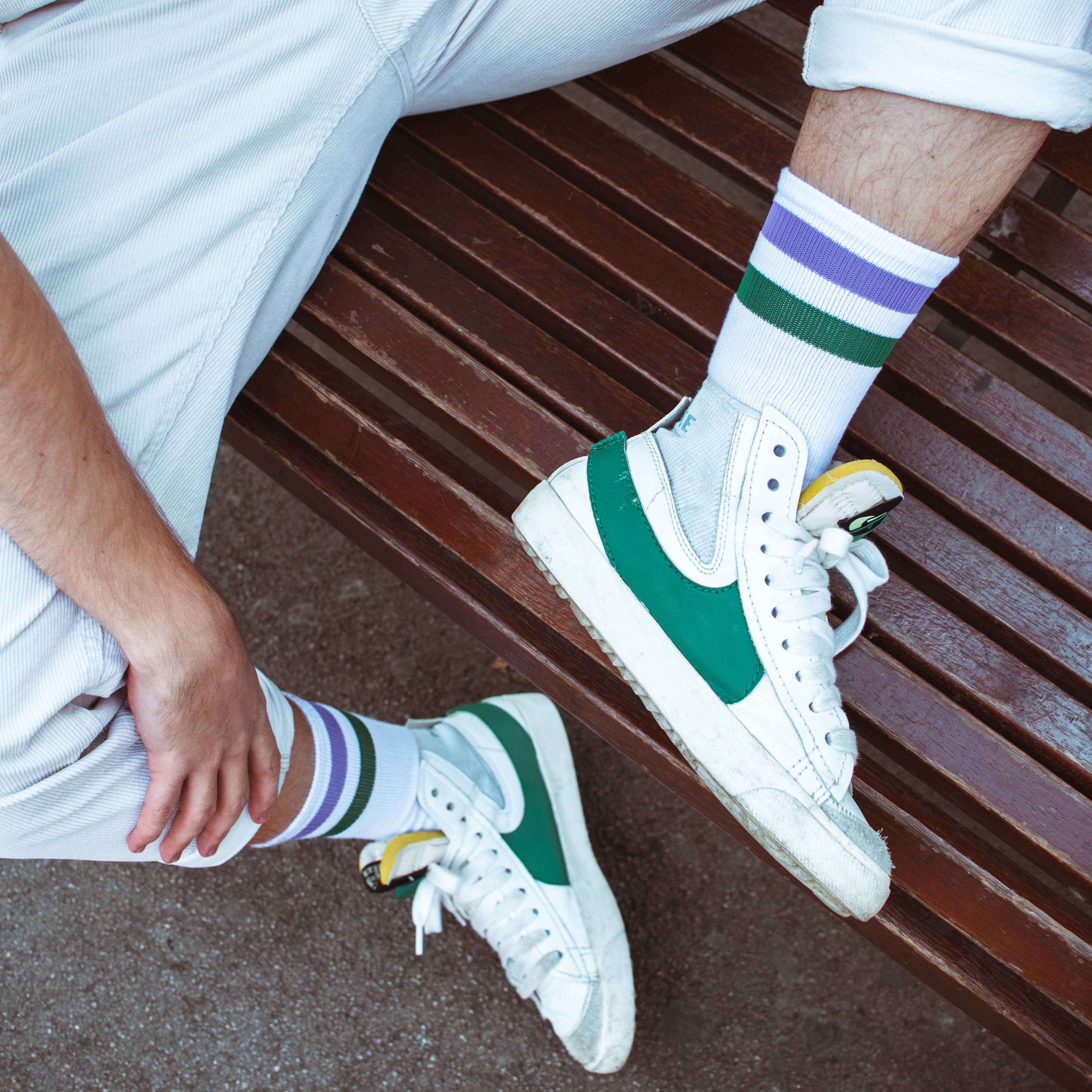 American Socks Joker Mid High Person auf Bank mit American Socks Joker Mid High, weißer Hose und Turnschuhen mit grünem Detail. Die Socken zeigen Streifen in Lila und Grün.