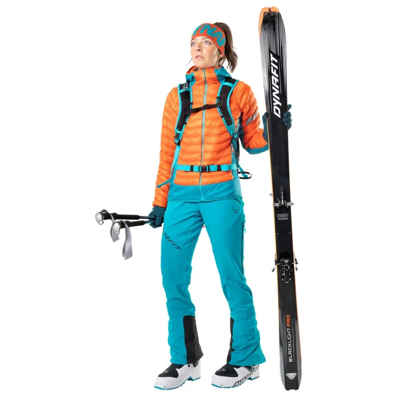 Femme en tenue de ski avec skis et bâtons. Elle porte un pantalon bleu pétrole Dynafit TLT Touring.
