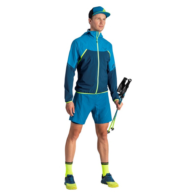 Mann in Sportkleidung: blaue Dynafit Alpine Pro Shorts, Jacke und Kappe. Er hält Wanderstöcke.