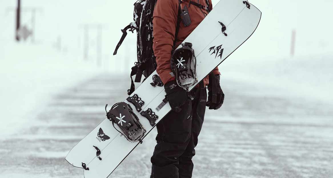 Une personne en tenue d'hiver tient un splitboard avec des fixations dans un environnement enneigé.