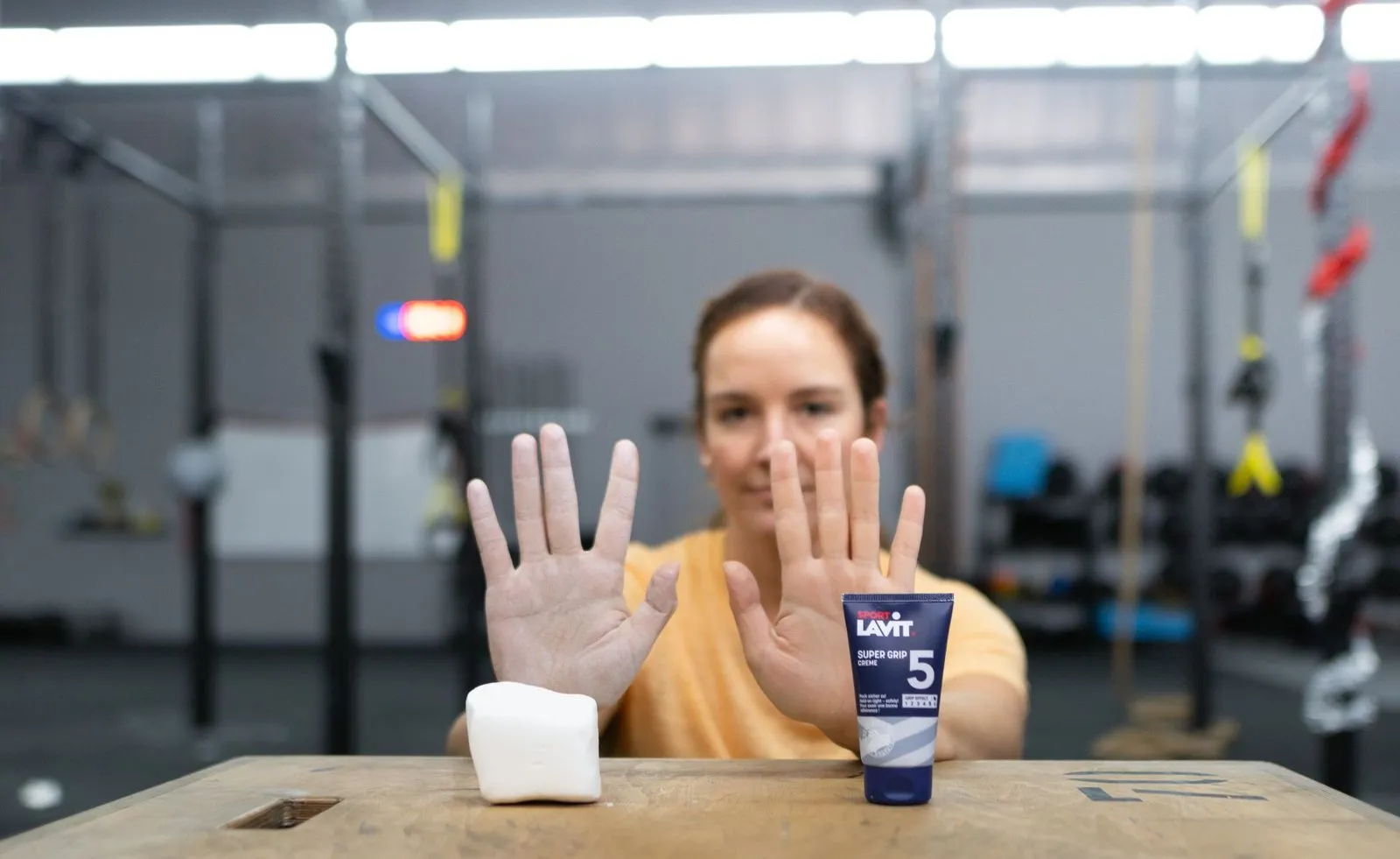 Donna che mostra le mani, il tubetto di crema Sport Lavit Super Grip e un blocco di magnesio su un tavolo di legno in palestra.
