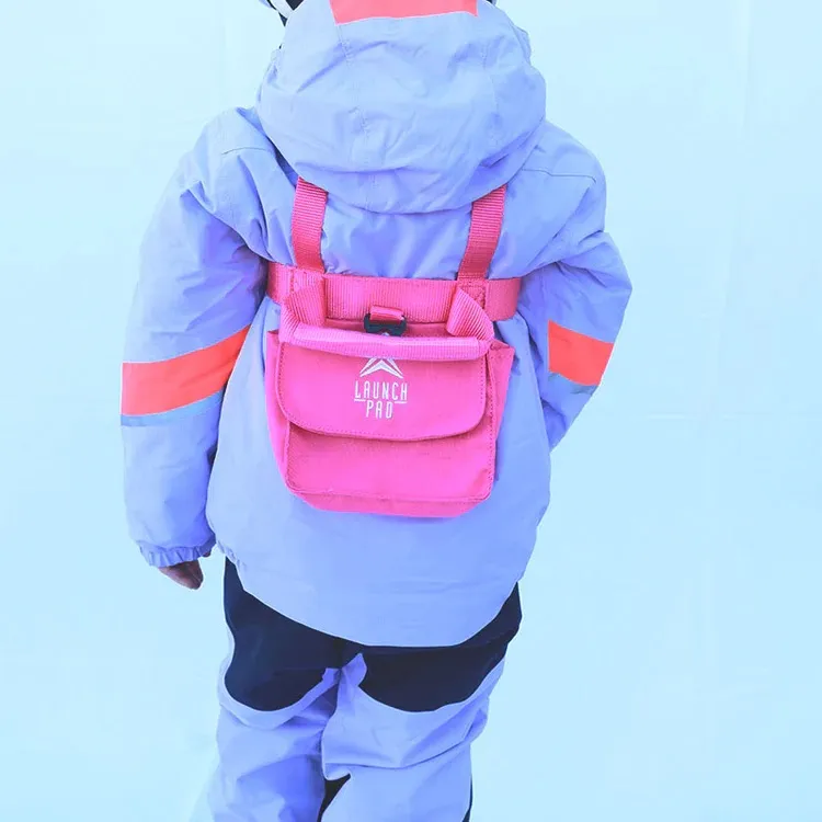 From behind: Toddler in a lavender-colored ski jacket and pants with bright orange stripes on the sleeves. On the back, a pink ski harness with a pouch and a white “Launch Pad” logo. Straps across the shoulders and waist.
