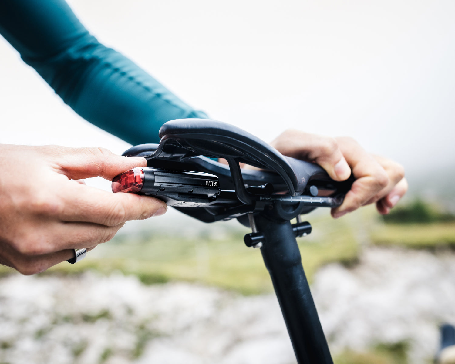 A person attaches a red rear bicycle light to a black bicycle saddle. A blurred landscape can be seen in the background.
