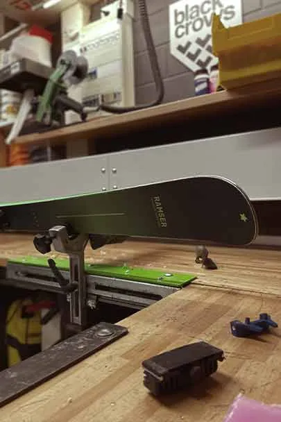 A ski is fixed in a vice on a workbench, surrounded by tools.