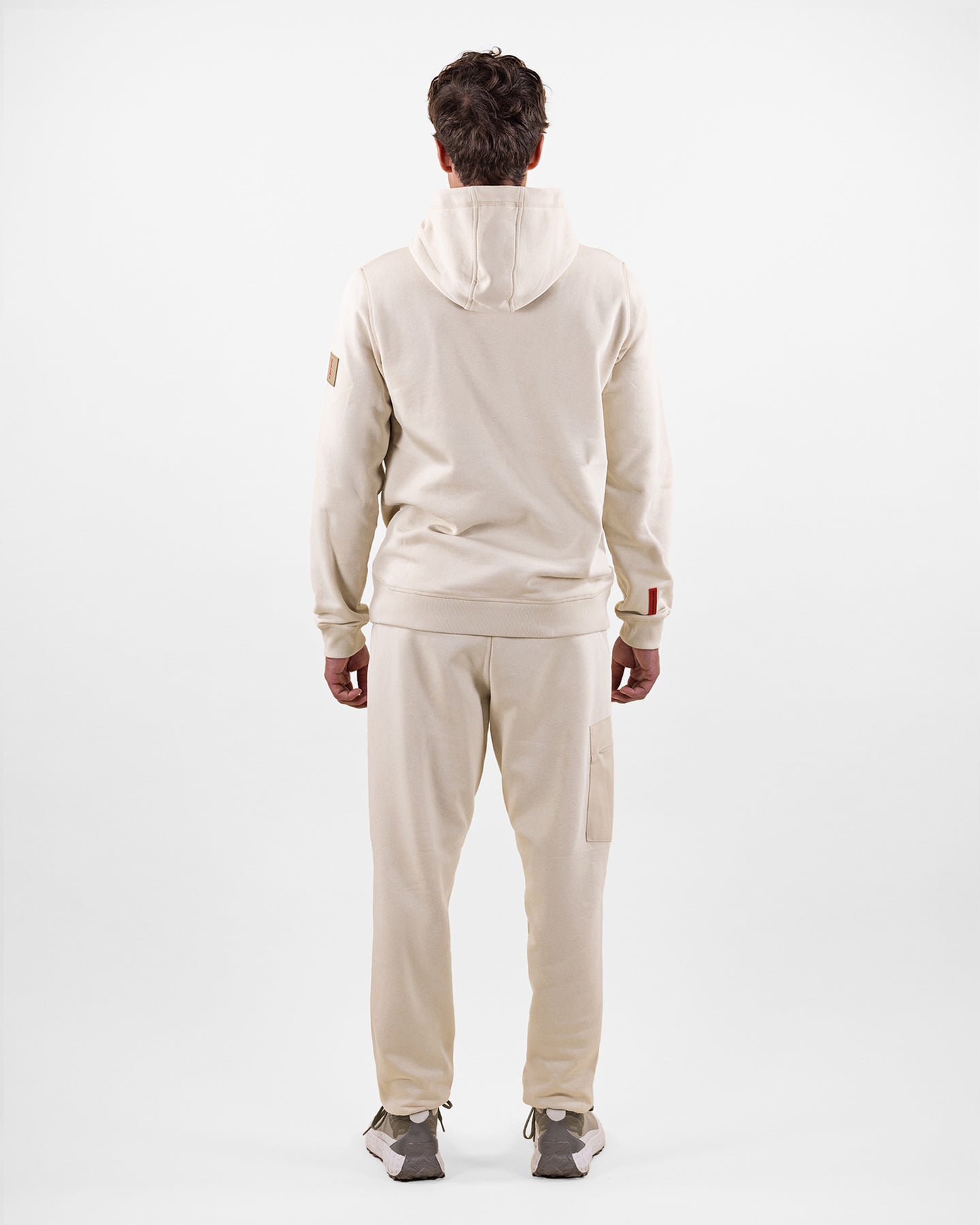 Vista posteriore di un uomo in tuta beige con cappuccio e scarpe da ginnastica, su sfondo bianco.