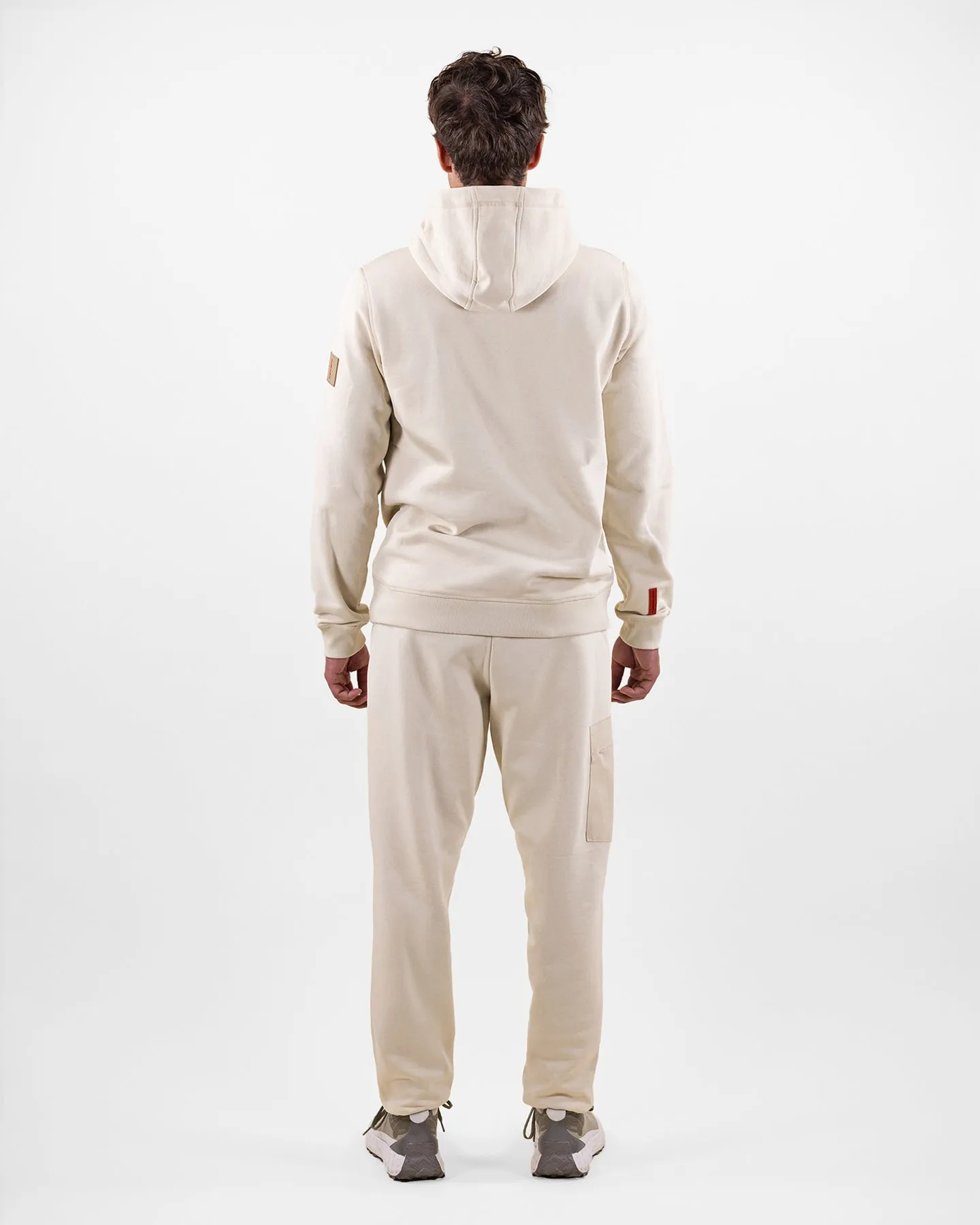 Vista posteriore di un uomo in tuta beige con cappuccio e scarpe da ginnastica, su sfondo bianco.