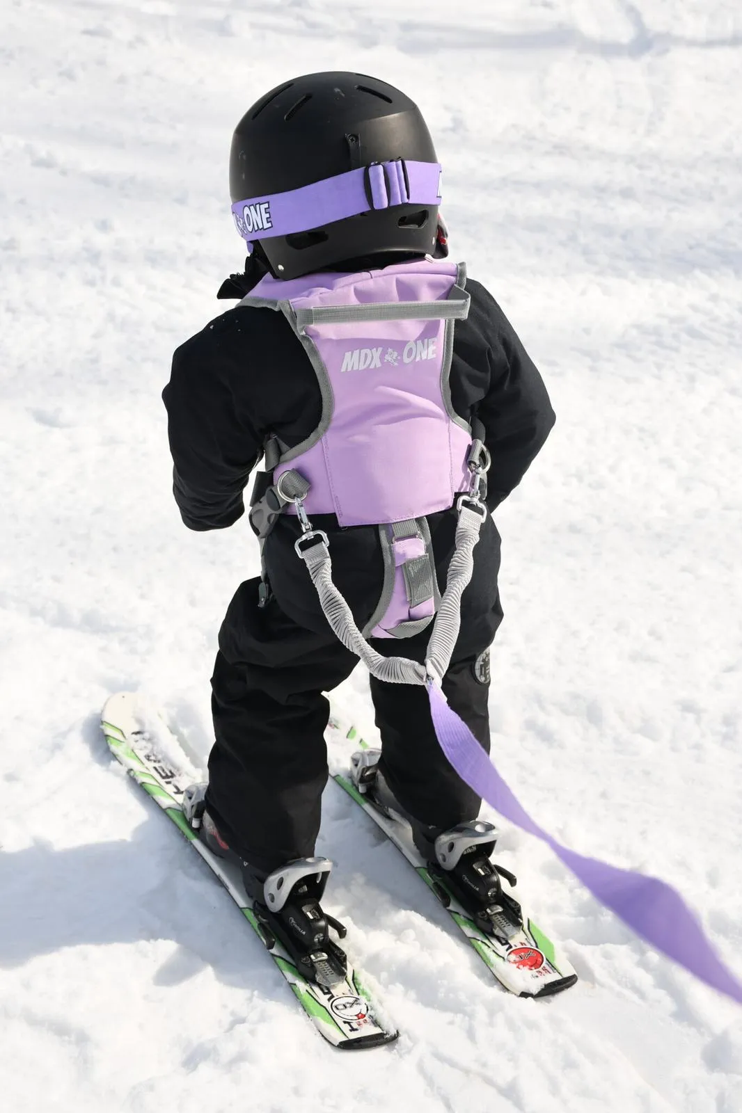 Kind mit Skiausrüstung, schwarzem Helm, lila Gurtzeug und Leine, steht auf Schnee.