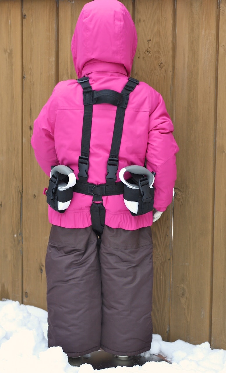 L'enfant avec une veste rose et un pantalon marron porte Lil' Ripper Gripper Aide au ski avec des cordes au dos.