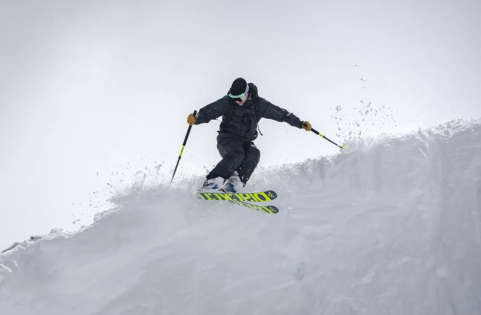 Skier in powder snow with Black Crows Atris skis in action, poles inserted.