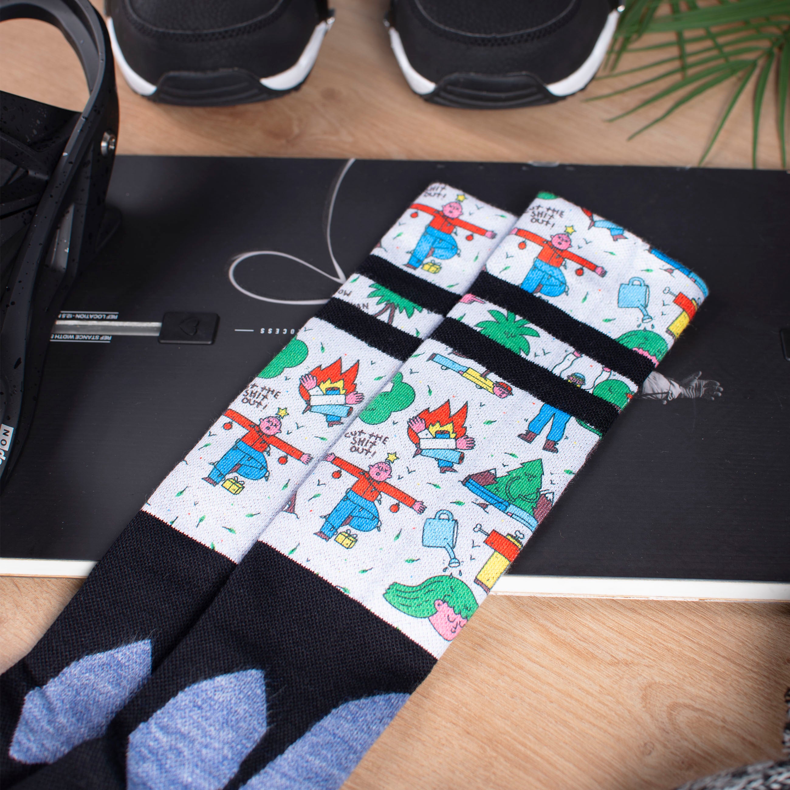 Comic Snow Socks Kniehohe Snow Socks auf Snowboard, Motiv mit Comicfiguren, schwarz-weiß gestreift, dazu Snowboard Boots und ein Blatt.