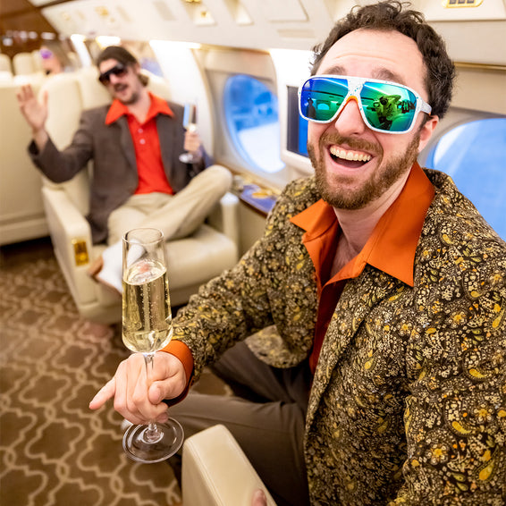 Lachender Mann mit South Beach Flight Optics Brille im Flugzeug, Sektglas in der Hand. Im Hintergrund eine weitere Person.