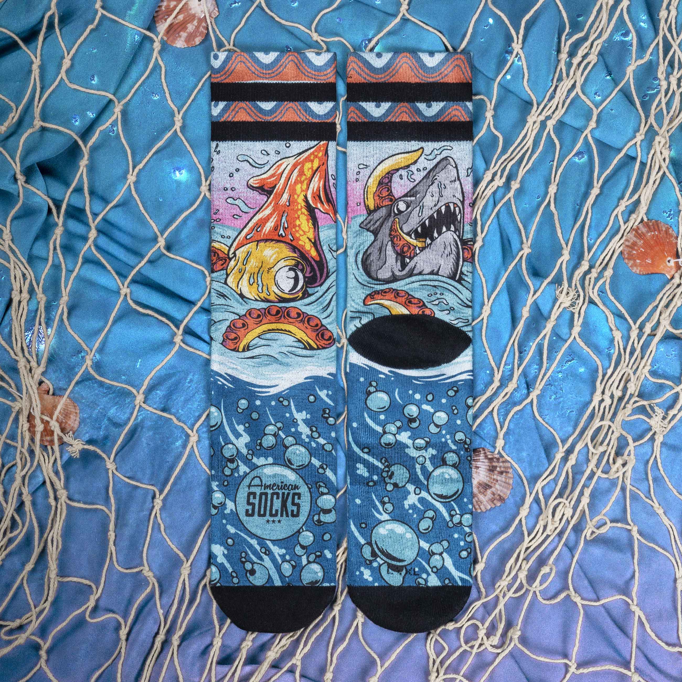 Seamonster Socken Blau Zwei 'Seamonsters Mid High' Socken mit Tintenfisch, Hai, Blasen und 'American Socks' Logo auf blauem Stoff mit Muscheln und Fischernetz.