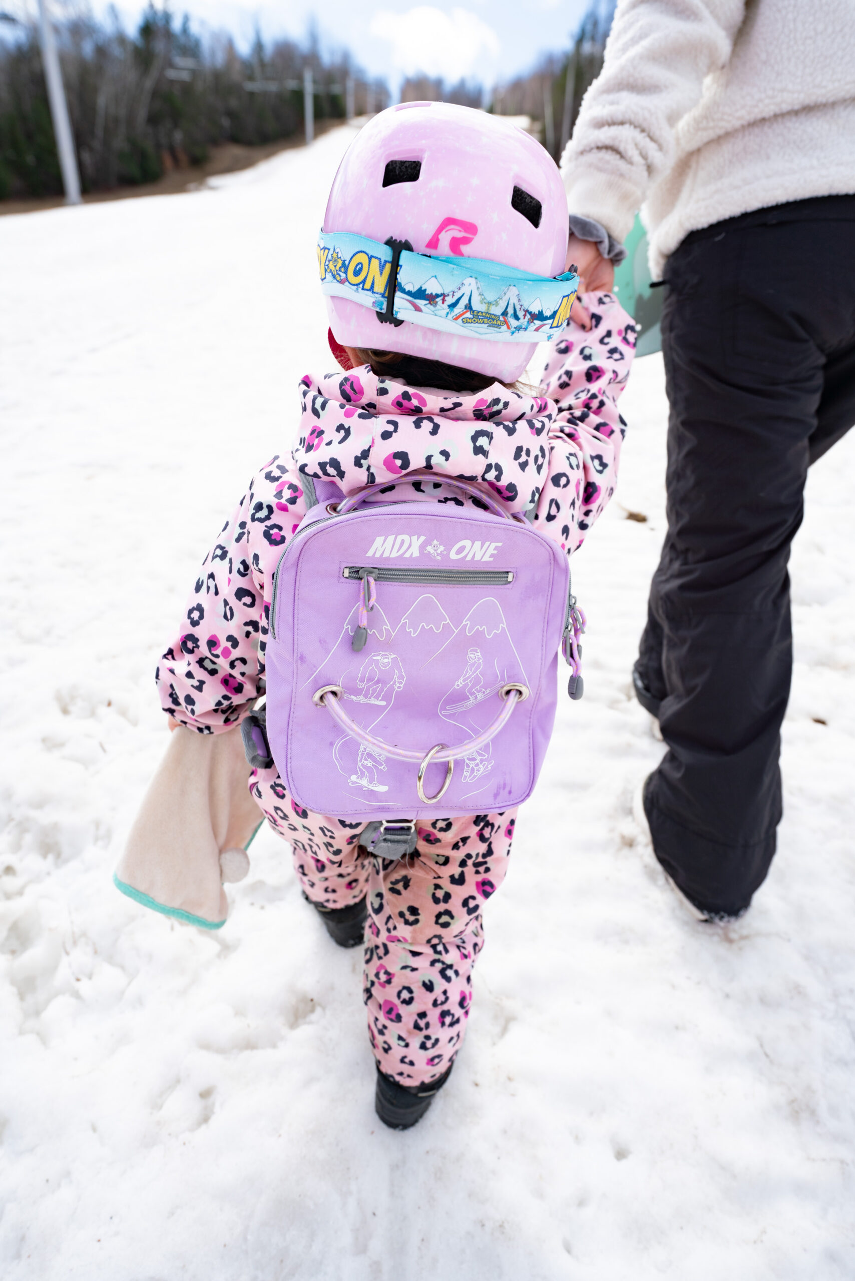 Un petit enfant en combinaison de ski rose léopard, casque rose et lunettes, descend une piste enneigée. Il porte un dispositif d'apprentissage du snowboard violet « THE ONE » avec harnais dorsal, guidé par la main.