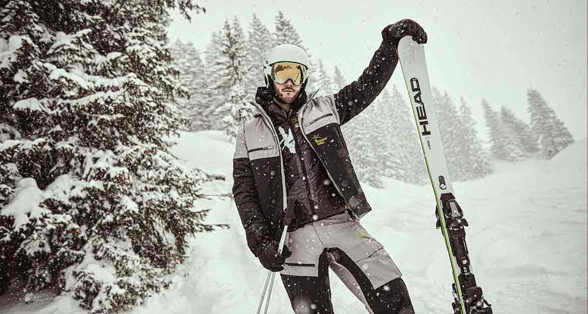 Un skieur en tenue d'hiver se tient devant des arbres enneigés et tient un ski.
