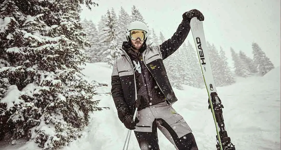 A skier in winter clothing stands in front of snow-covered trees and holds a ski.
