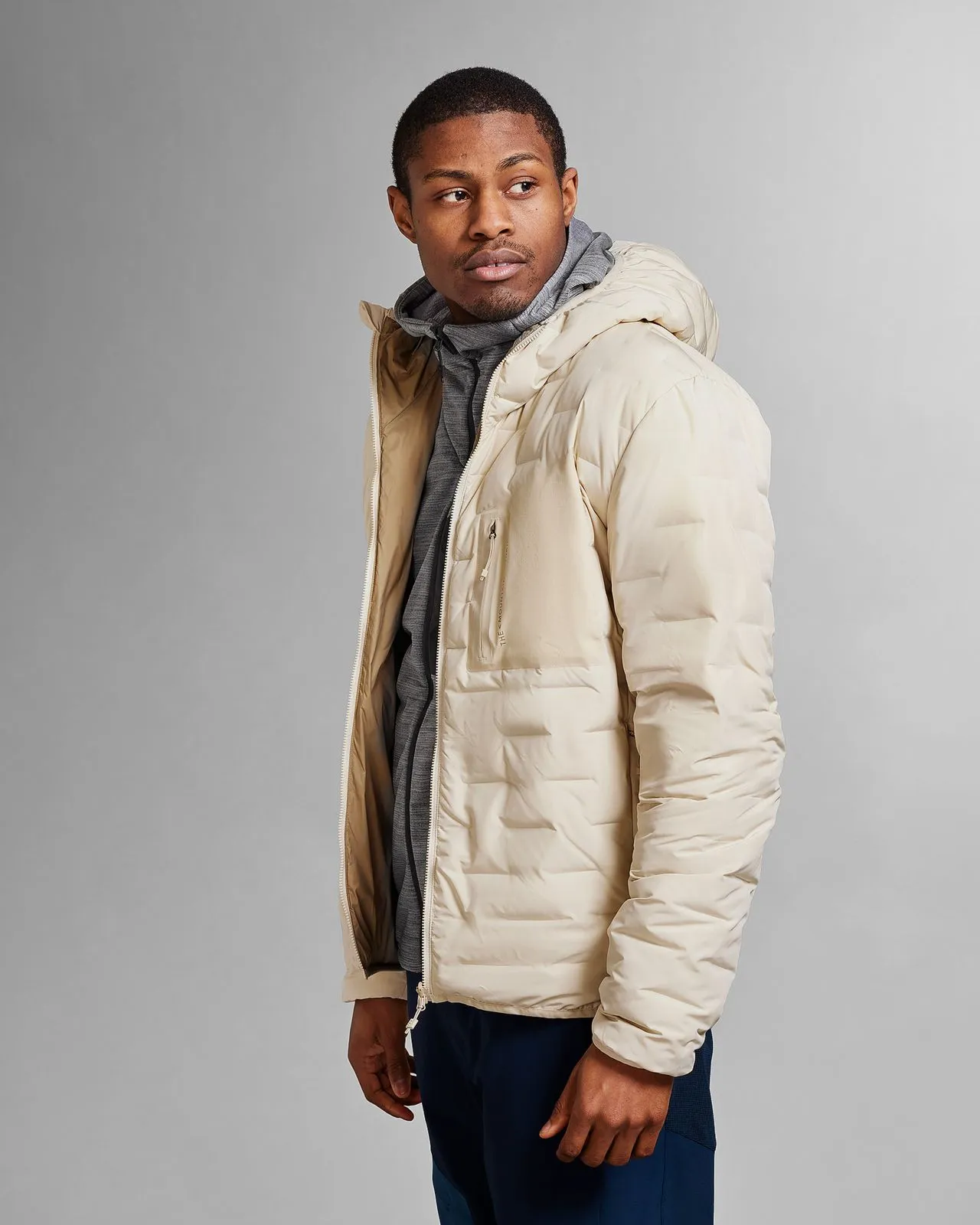 Man with "Castle Wall Sand" hooded jacket. Quilted jacket, zipper open, gray hoodie underneath. View from the side.