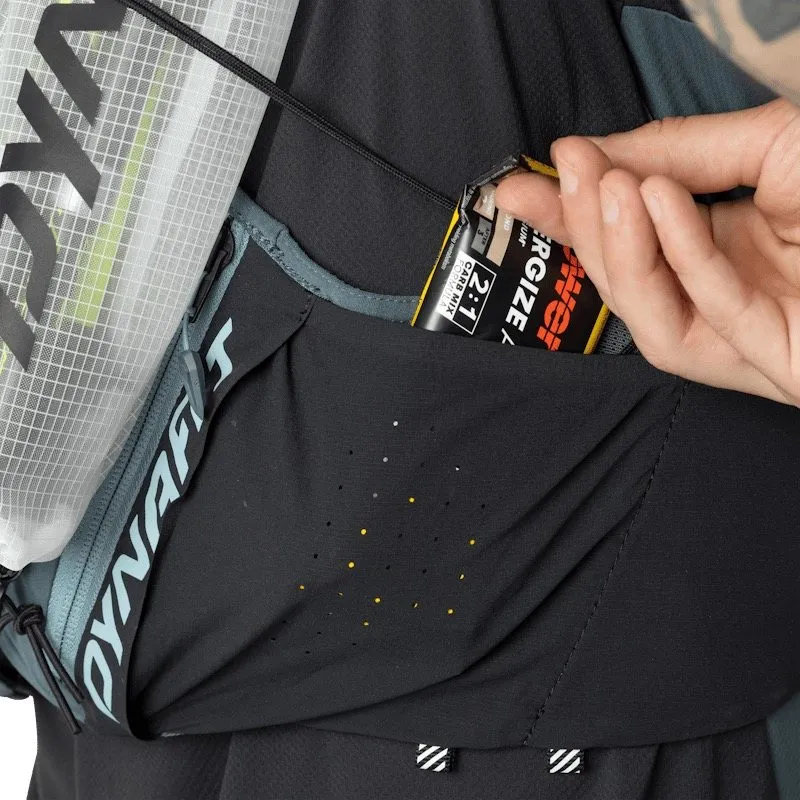 Close-up of a black Dynafit running backpack. A hand is taking a Powerbar energy bar out of a side pocket. The perforated pocket shows small yellow dots. A hydration bladder with a white mesh pattern is visible.