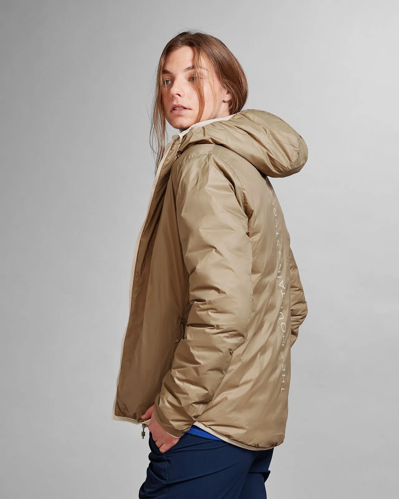 Person wearing the Reversible Light Hood Jacket in Castle Wall Sand, view over the shoulder. Text "Mountain Studio" visible.