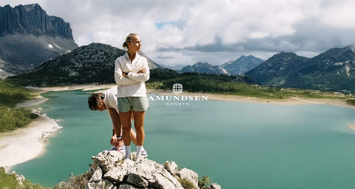 Zwei Personen auf Felsen mit Blick auf einen See. Eine Person bindet ihre Schuhe, die andere blickt in die Ferne. Bergige Landschaft im Hintergrund.