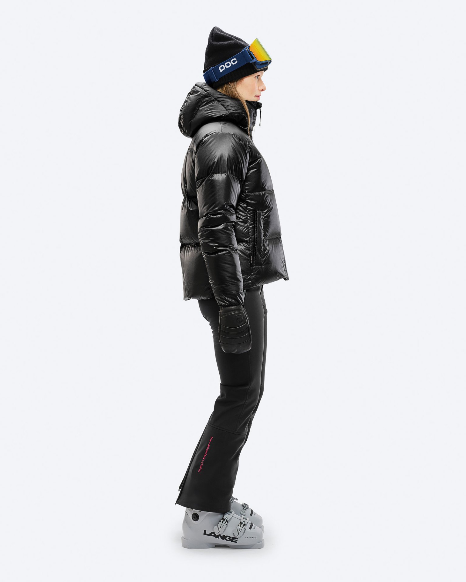 Woman skier in black Woman in black ski pants and jacket from the side, with ski boots, cap and ski goggles.