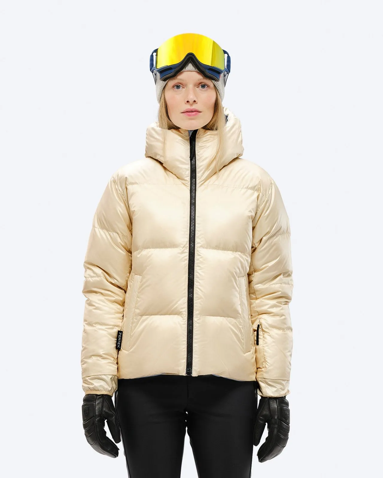 A woman wears a shiny beige down jacket with a hood, black gloves and ski goggles against a white background.