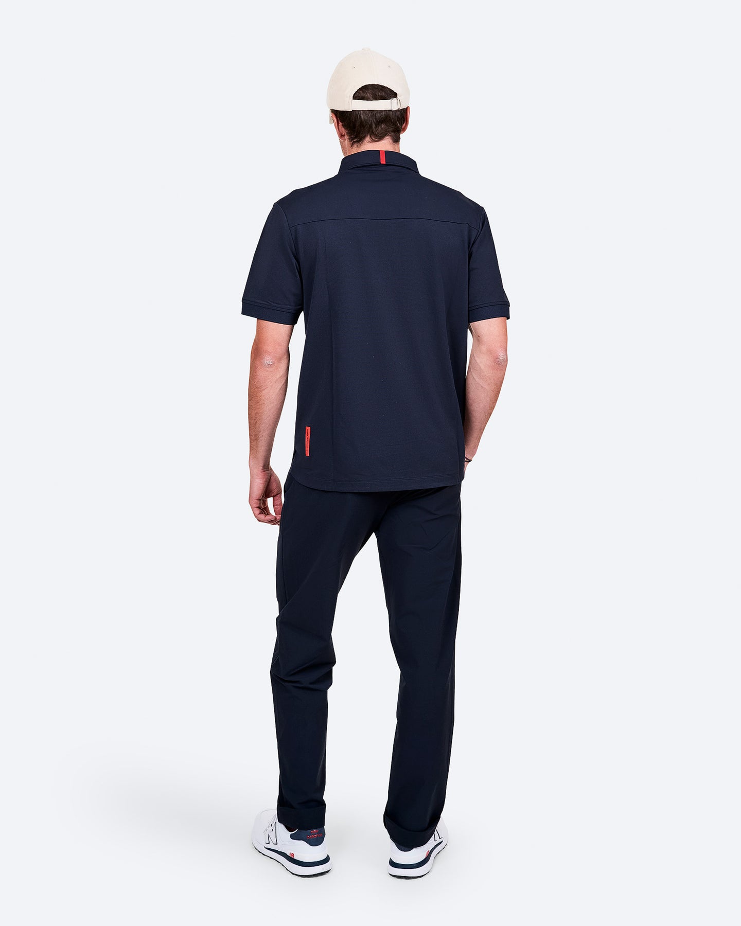 Vue de dos d'un homme vêtu de bleu marine, d'une casquette beige et de baskets blanches sur fond blanc.