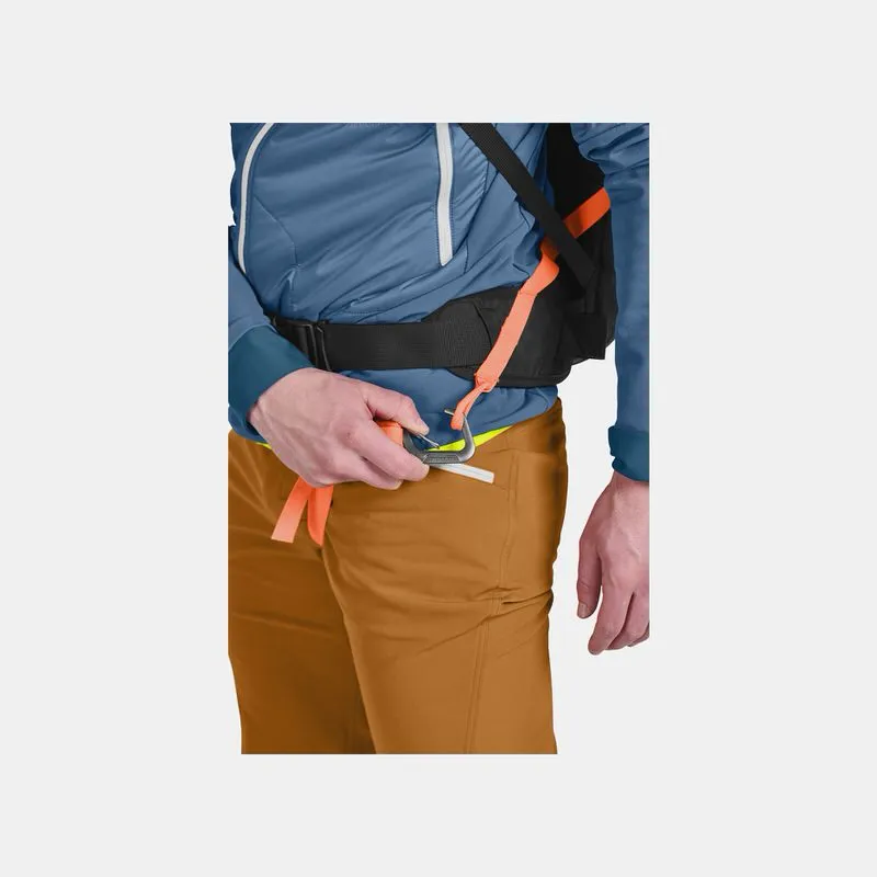 Detailaufnahme: Person befestigt Beingurt am Ortovox Avabag Litric Tour 30 Rucksack. Der Gurt ist orange, die Hose braun.