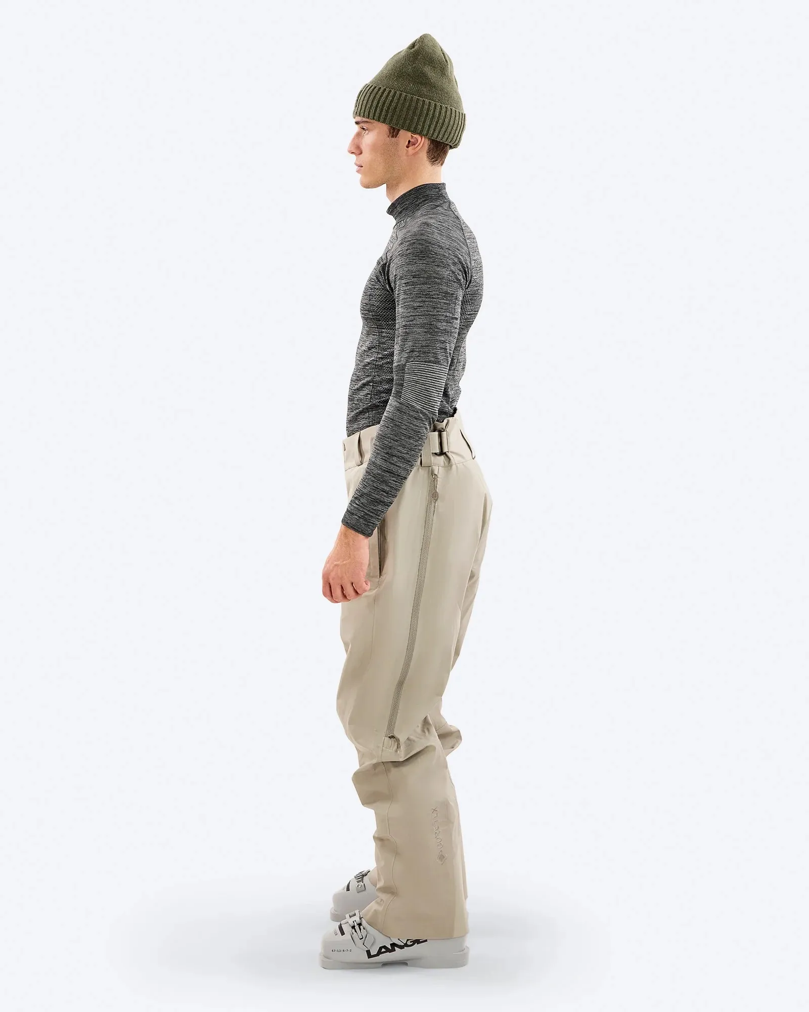 Man in ski outfit, side view. He wears a cap, tight top, ski pants and ski boots in front of a white background.