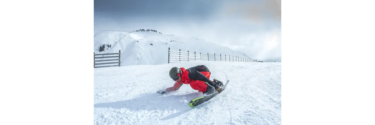 Un snowboarder en veste rouge carve dans la neige avec son snowboard Kizamu LYT. Paysage de montagne en arrière-plan.