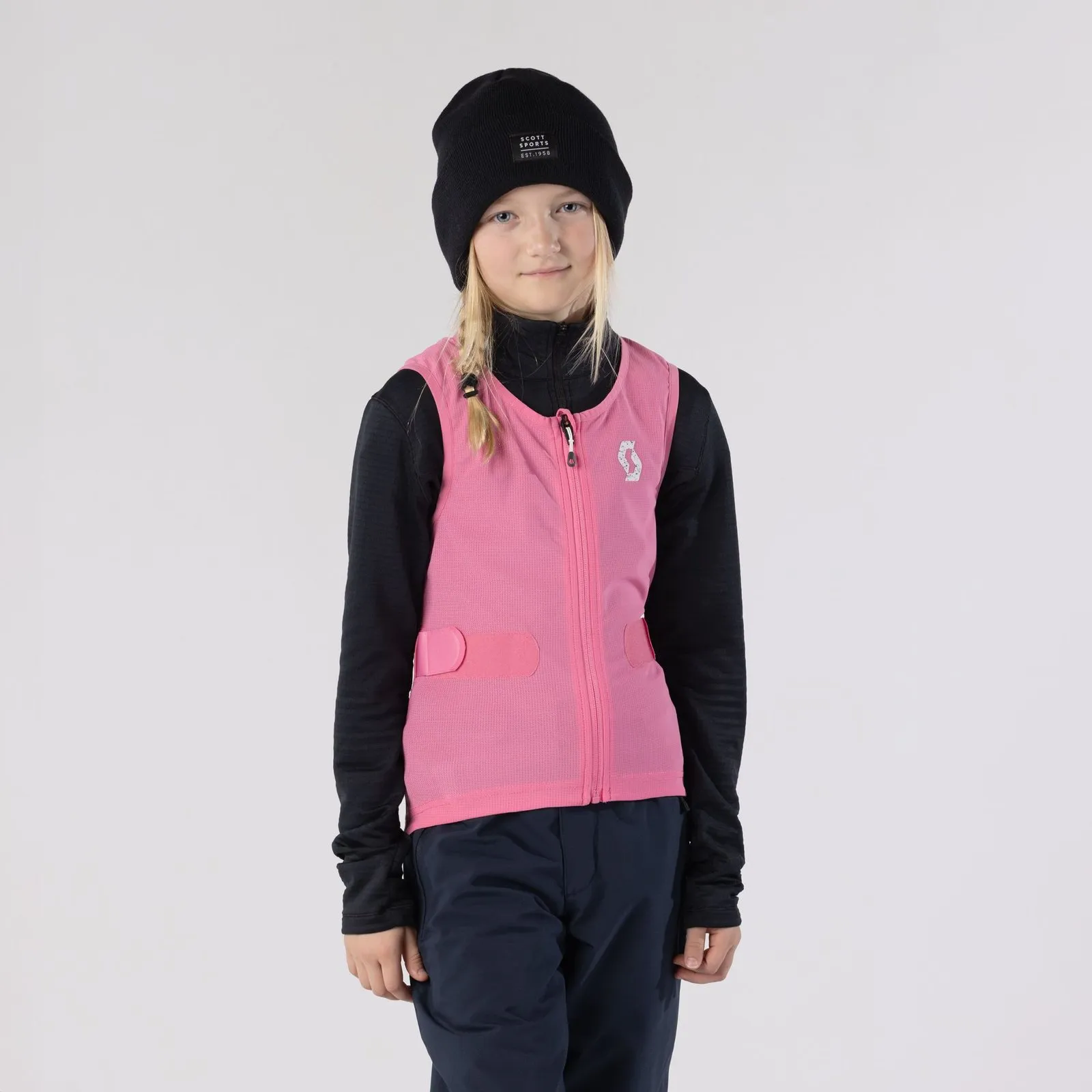 A young girl with blonde pigtails, black cap, black top and pink vest in front of a white background.