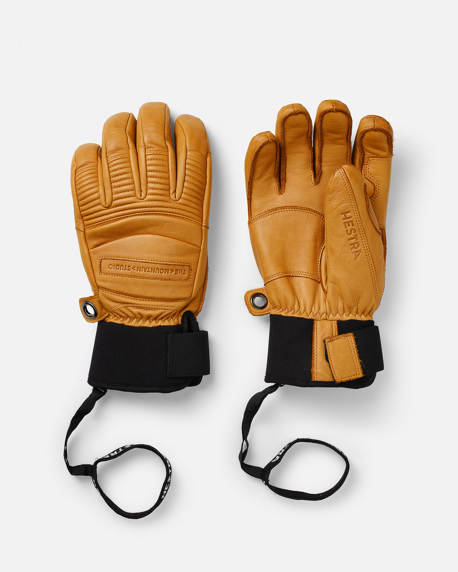 Sand-colored leather gloves Two Hestra® leather gloves in sand color with black neoprene cuffs and safety straps on a white background.