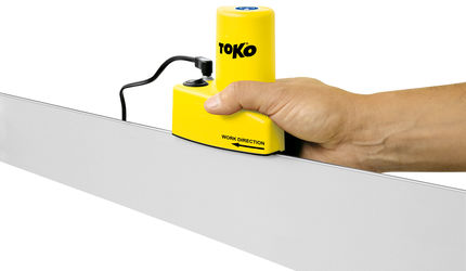 Appareil de réglage des carres de ski La main guide le Toko Edge Tuner jaune sur la carre du ski. L'affûteuse a un câble d'alimentation. Flèche "Work Direction" sur l'appareil.