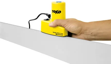 Hand führt gelben Toko Edge Tuner über Skikante. Das Schleifgerät hat ein Stromkabel. Pfeil "Work Direction" auf dem Gerät.