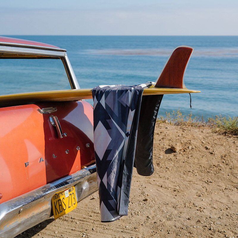 Rumpl Shammy Towel im Woven Daydream Design hängt auf Surfbrett an einem roten Auto am Strand.