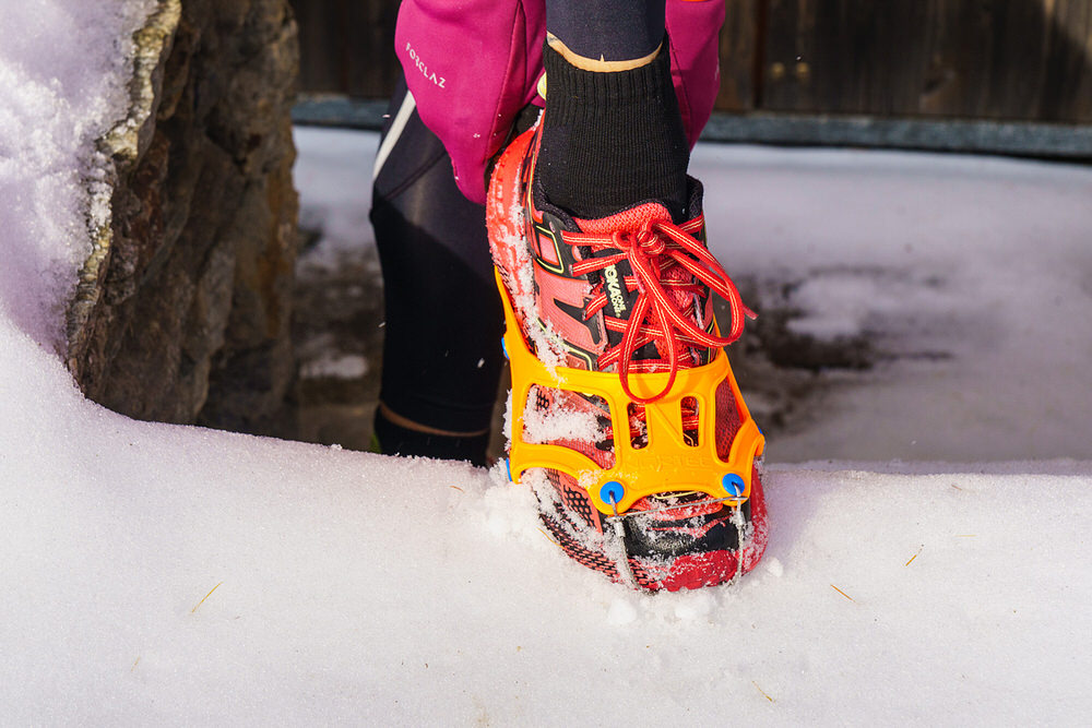 Gros plan sur une chaussure de trail running avec une griffe jaune sur la neige. Les pointes en acier de la griffe assurent le maintien.