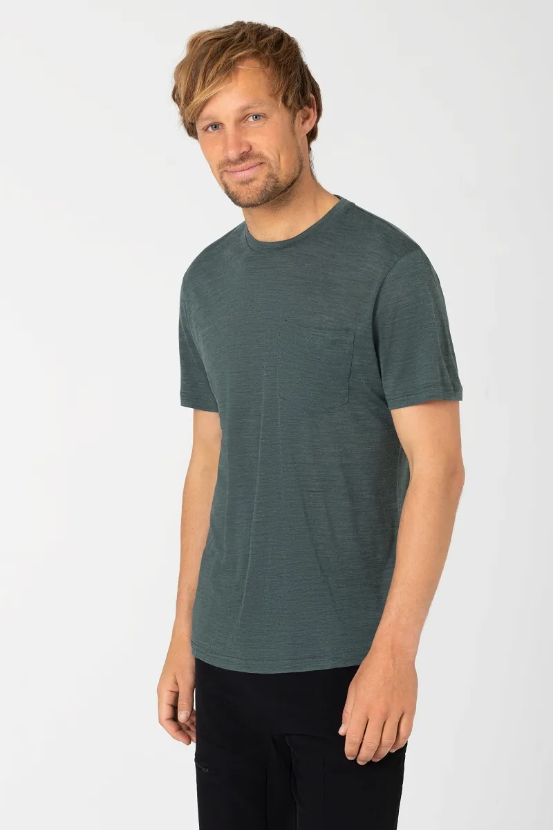 Uomo con maglietta color petrolio screziata con taschino e pantaloni neri. Casual, guarda di lato.