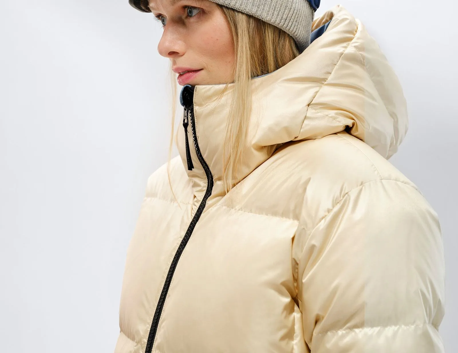 Profile of a woman wearing a beige down jacket and hat. The jacket has a black zipper and a hood.